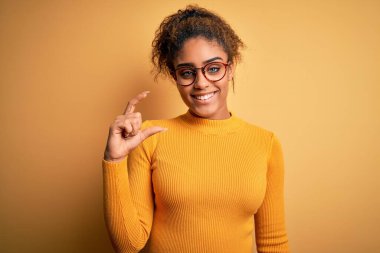 Sarı arka planda süveter ve gözlük takan güzel afro-amerikalı kız gülümsüyor ve el hareketleriyle küçük beden işareti yaparak kameraya bakıyor. Ölçü kavramı.