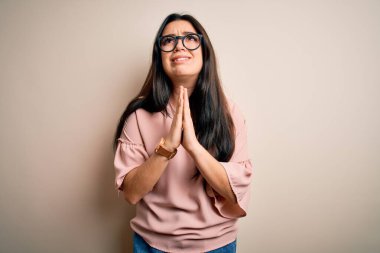 Soyutlanmış arka planda gözlük takan genç, zarif bir kadın elleri bağlı yalvarıyor ve dua ediyor. Yüzünde umut ifadesi var. Çok duygusal ve endişeli. Yalvarıyorum..