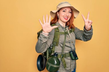 Sırt çantalı, sarı şapkalı, sırt çantalı, genç, kızıl saçlı kadın, yedinci parmaklarını gösterirken kendine güvenen ve mutlu gülümseyen..