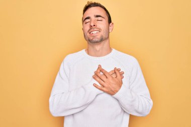 Mavi gözlü, sıradan bir süveter giyen, sarı arka planda duran, elleri göğsünde, gözleri kapalı ve yüzünde minnet ifadesi olan genç yakışıklı bir adam. Sağlık kavramı.