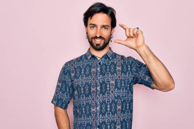 Genç, İspanyol asıllı bohem bir adam pembe arka planda hippi tarzı giyinmiş, gülümseyen ve kendine güvenen, el hareketleriyle küçük boy işareti yapan parmaklarıyla kameraya bakan. Ölçü kavramı.