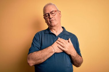 Orta yaşlı yakışıklı beyaz adam sarı arka planda günlük polo ve gözlük takıyor elleri göğsünde, gözleri kapalı ve yüzünde minnet dolu bir jest ile gülümsüyor. Sağlık kavramı.