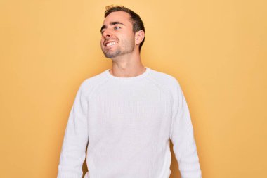 Mavi gözlü, sıradan bir süveter giyen, sarı arka planda dikilen, yüzünde gülümseme, doğal bir ifade olan genç yakışıklı bir adam. Kendinden emin gülen.