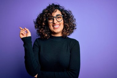 Genç ve güzel kıvırcık Arap kadın, mor arka planda günlük süveter ve gözlük takıyor. Yüzünde kocaman bir gülümseme, eli ve parmağı kameraya bakıyor..