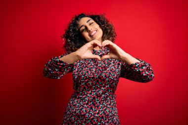 Genç, güzel, kıvırcık, çiçek desenli elbiseler giyen, kırmızı arka planda dikilen, aşık olmuş, elleriyle kalp sembolü yapan bir kadın. Romantik konsept.