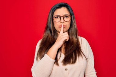 Kırmızı izole edilmiş arka planda duran gözlüklü, genç, Latin kökenli, zeki bir kadın sessiz olmak istiyor. Sessizlik ve gizli konsept.