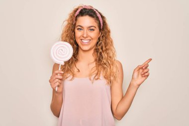 Mavi gözlü, güzel, sarışın bir kadın pembe arka planda şeker yiyerek mutlu bir şekilde elini ve parmağını kenarı işaret ediyor.