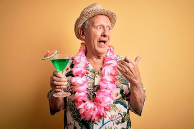 Beyaz saçlı, yaz şapkası takan ve Hawaii kolyesi takan kokteyl içen ve gülümseyen bir yüzle baş parmağıyla kendini gösteren yaşlı bir adam.