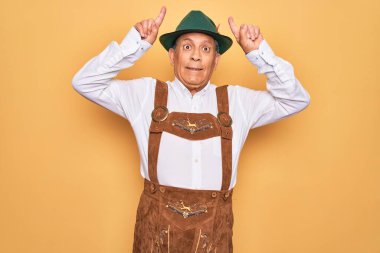 Sarı arka planda Alman geleneksel Ekim Festivali kıyafeti giyen yaşlı gri saçlı adam boynuzları gibi parmağıyla komik hareketler yapıyor.