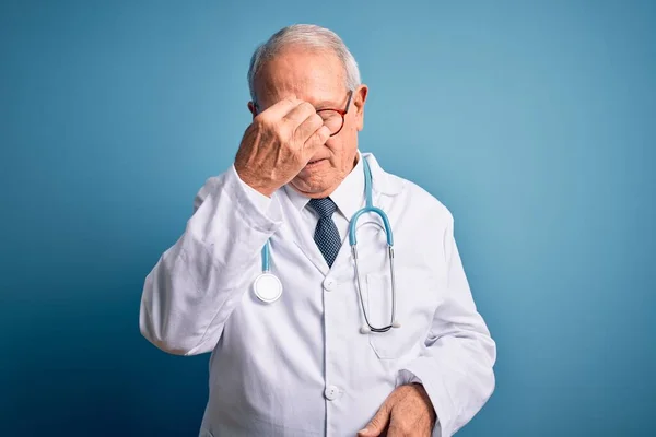 青い背景色の上に聴診器と医療コートを身に着けているシニア灰色の髪の医者の男は 鼻や目をこすり疲れや頭痛を感じ疲れ ストレスとフラストレーションの概念 — ストック写真