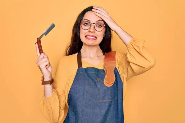Young beautiful barber woman wearing apron holding razor blade over yellow background stressed with hand on head, shocked with shame and surprise face, angry and frustrated. Fear and upset for mistake.