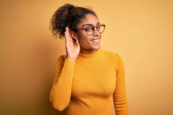 黄色の背景にセーターと眼鏡を身に着けている若い美しいアフリカ系アメリカ人の女の子は 噂やゴシップへの聴聞会を聞いて手で笑っています 病気の概念 — ストック写真