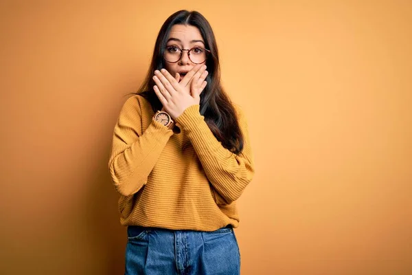 若いブルネットの女性は間違いのために手で口をカバーショックを受けた黄色の隔離された背景に眼鏡とカジュアルなセーターを着て 秘密概念 — ストック写真