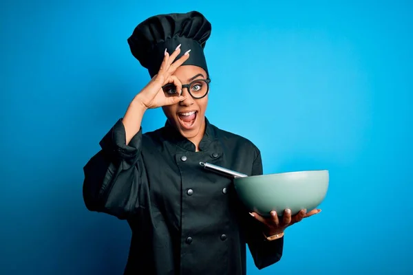 Young african american chef girl wearing cooker uniform and hat using whisk and bowl with happy face smiling doing ok sign with hand on eye looking through fingers