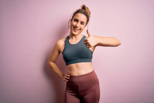 Young beautiful blonde sportswoman doing sport wearing sportswear over pink background doing happy thumbs up gesture with hand. Approving expression looking at the camera showing success.