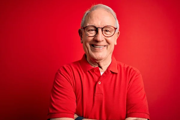 Grey haired senior man wearing glasses and casual t-shirt over red background happy face smiling with crossed arms looking at the camera. Positive person.