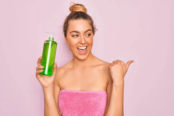 Beautiful blonde woman with blue eyes wearing towel shower after bath holding aloe vera gel pointing and showing with thumb up to the side with happy face smiling