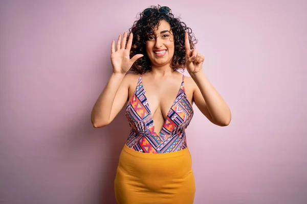 Young beautiful arab woman on vacation wearing swimsuit and sunglasses over pink background showing and pointing up with fingers number six while smiling confident and happy.
