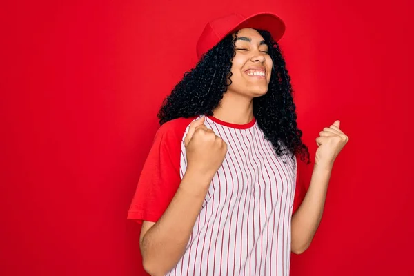 Young african american curly sportswoman wearing baseball cap and striped t-shirt excited for success with arms raised and eyes closed celebrating victory smiling. Winner concept.