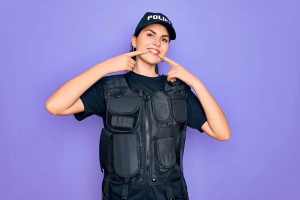 Young police woman wearing security bulletproof vest uniform over purple background smiling cheerful showing and pointing with fingers teeth and mouth. Dental health concept.