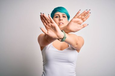 Mavi saçlı, beyaz arkaplan üzerine günlük tişört giyen genç ve güzel bir kadın. Reddedilmiş yüz ifadesi. Kollarını kavuşturup, avuçlarını ters çevirip, kızgın yüz ifadesi takınıyor.