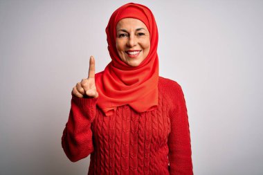 Orta yaşlı esmer kadın izole edilmiş beyaz arka plan üzerine geleneksel başörtüsü takıyor ve bir numaralı parmağını gösterirken kendinden emin ve mutlu gülümsüyor..