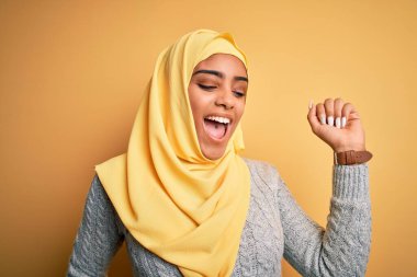 Genç, güzel, Afro-Amerikalı bir kız, izole edilmiş sarı arka planda Müslüman başörtüsü takıyor mutlu ve neşeli dans ediyor, hareketli, rahat ve kendinden emin bir şekilde müzik dinliyor.