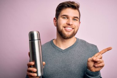 Sakallı ve mavi gözlü genç bir adam pembe zemin üzerinde su olan termosu tutuyor.