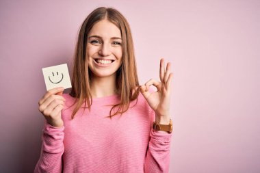 Genç, güzel, kızıl saçlı kadın elinde gülümseyen emoji mesajıyla hatırlatma kağıdı tutuyor. Tamam işareti yapıyor. Mükemmel bir sembol.