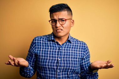 Sarı arka plan üzerine gündelik tişört ve gözlük takan genç, yakışıklı Latin bir adam. Hiçbir fikri olmayan, kolları açık bir adam..