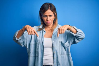 Orta yaşlı, güzel bir kadın, yalnız mavi arka planda dikilmiş üzgün ve üzgün görünüyor, parmaklarıyla yönü gösteriyor, mutsuz ve depresif..