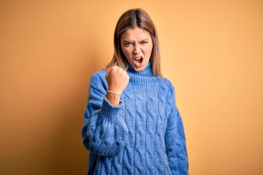 Sarı arka plan üzerine balıkçı yaka kazak giyen genç ve güzel bir kadın öfkeyle bağırırken kızgın ve kızgın bir şekilde yumruk kaldırıyor. Öfke ve agresif kavram.