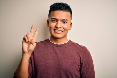 İzole edilmiş beyaz arka planda dikilen ve kameraya gülümseyen, zafer işareti yapan, gündelik süveter giyen genç, yakışıklı Latin adam. İki numara..