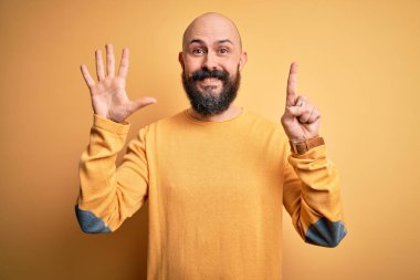 Sarı arka planda dikilen sıradan süveter giyen sakallı yakışıklı kel adam kendini beğenmiş ve mutlu bir şekilde gülümserken altı numarayı işaret ediyor..