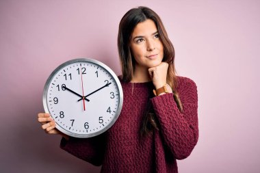 Geri sayım yapan genç güzel bir kız büyük bir saati soyutlanmış pembe arka plan üzerinde tutuyor ciddi bir yüz soru düşünüyor, çok karışık bir fikir.