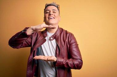 Sarı arka plan üzerinde moda deri ceket ve güneş gözlüğü takan genç, yakışıklı ve modern bir adam. Büyük ve büyük boyut işareti gösteren el hareketleriyle, ölçü sembolü. Kameraya bakıp gülümsüyor. Ölçüm konsepti.
