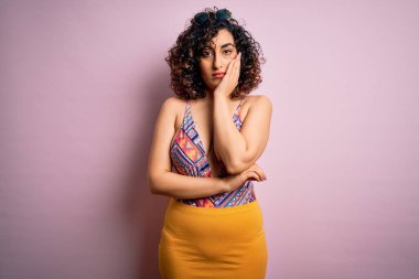 Genç, güzel bir Arap kadın, mayo ve pembe arka planda güneş gözlüğü takarak tatilde. Kollarını kavuşturup depresyondan sıkılmış ve yorgun görünüyor..