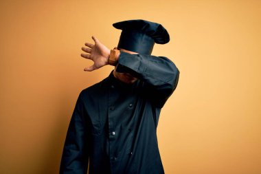 Aşçı üniforması giyen genç Brezilyalı şef ve izole edilmiş sarı arka planı olan şapkalı adam kollarıyla gözlerini kapatıyor, ciddi ve üzgün görünüyor. Görme, saklama ve reddetme konsepti