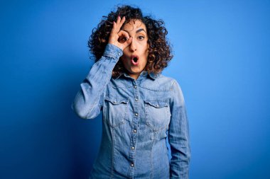 Genç ve güzel kıvırcık Arap kadın mavi arka plan üzerinde sıradan bir kot gömlek giyiyor. Şaşırmış yüz ifadesiyle şaşırmış, parmaklarının arasından bakıyor. İnançsız ifade.