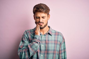 Sakallı genç yakışıklı bir adam pembe arka planda dikilmiş diş ağrısı ya da diş hastalığından dolayı acı dolu bir ifadeyle ağıza dokunuyor. Dişçi