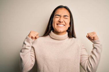 Genç, güzel Asyalı bir kadın beyaz arka plan üzerine balıkçı yaka kazak giyiyor. Çok mutlu ve heyecanlı. Kolları havada, gülümseyerek ve başarı için çığlık atarak kazanan jestler yapıyor. Kutlama konsepti.
