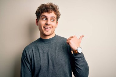 Sarışın, kıvırcık saçlı, beyaz arka planda sıradan bir süveter giyen, gülümseyen ve baş parmağıyla yüzünü gösteren genç, yakışıklı bir adam..