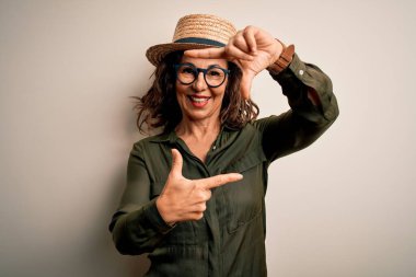 Orta yaşlı esmer kadın gözlüklü ve şapkalı izole edilmiş beyaz arka planda gülümseyen elleri ve parmakları mutlu yüzlü olan bir çerçeve yapıyor. Yaratıcılık ve fotoğrafçılık kavramı.