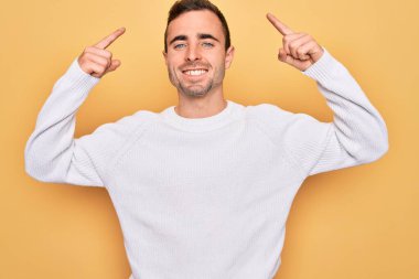 Mavi gözlü, sıradan bir süveter giyen, sarı arka planda durup gülümseyen, iki eliyle başını gösteren yakışıklı bir adam. Harika bir fikir ya da düşünce, iyi bir hafıza.