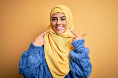 Genç, güzel, esmer Müslüman kadın izole edilmiş sarı arka planda Arap başörtüsü takıyor ağzı açık gülümsüyor, parmakları işaret ediyor ve zorla neşeli bir gülümseme sergiliyor.