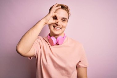 Genç, yakışıklı, kızıl saçlı, müzik dinleyen, pembe arka planda kulaklık takan, el hareketleriyle gülümseyen, mutlu yüzlü parmaklarından bakan bir adam..