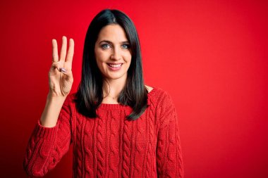 Mavi gözlü genç esmer kadın izole edilmiş kırmızı arka plan üzerine gündelik süveter giyiyor ve üç numarayı gösterirken kendinden emin ve mutlu bir şekilde gülümsüyor..