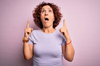 Orta yaşlı, kıvırcık saçlı, rahat bir tişört giyen güzel bir kadın pembe arka planda şaşkın şaşkın bakıyor, parmaklarıyla işaret ediyor ve kollarını kaldırıyordu..
