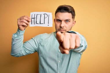 Mavi gözlü genç iş adamı sarı arkaplan üzerine kağıt tutarak yardım istiyor. Kamerayı işaret ediyor ve sen, el işareti, ön taraftan pozitif ve kendinden emin bir hareket.
