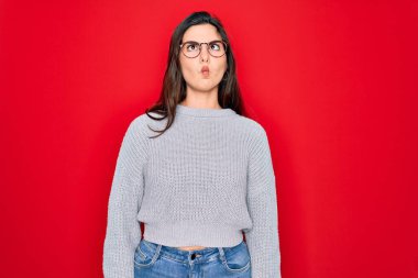 Genç, güzel esmer kadın kırmızı arka plan üzerine gündelik kazak giyip dudaklarıyla balık suratı yapıyor, çılgınca ve komik hareketler yapıyor. Komik ifade.